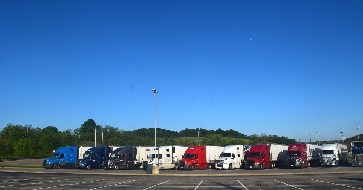 A row of semi-trucks parked at a U.S. fleet lot under a clear blue sky